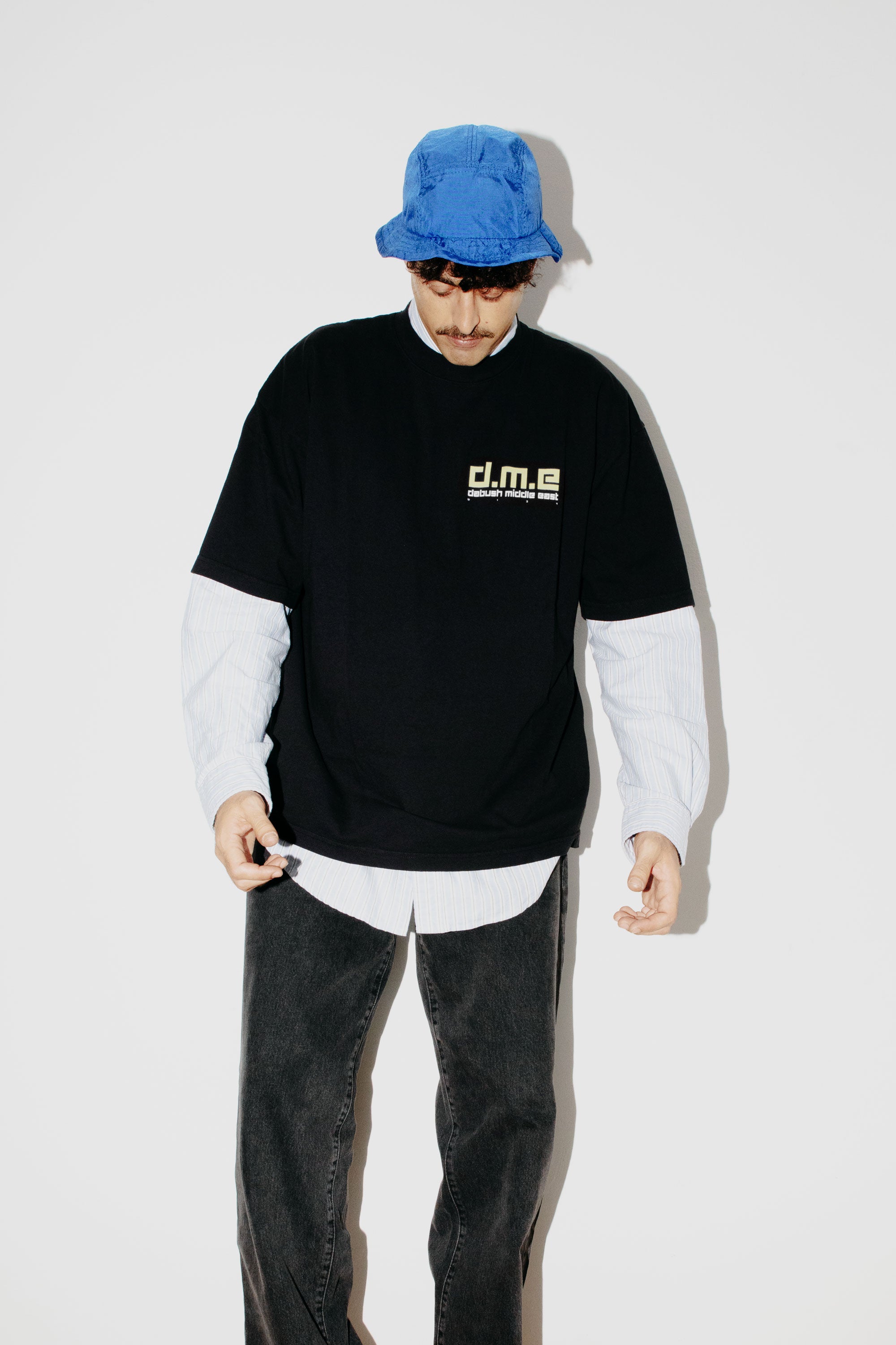 A model wearing an oversized DABUSH t-shirt in black, made from 100% cotton, with printed graphics Dabush Middle East.
דוגמן לובש אוברסייז טישרט של דבוש בצבע שחור, עשויה 100% כותנה עם גרפיקה ענקית מקדימה ומאחורה.