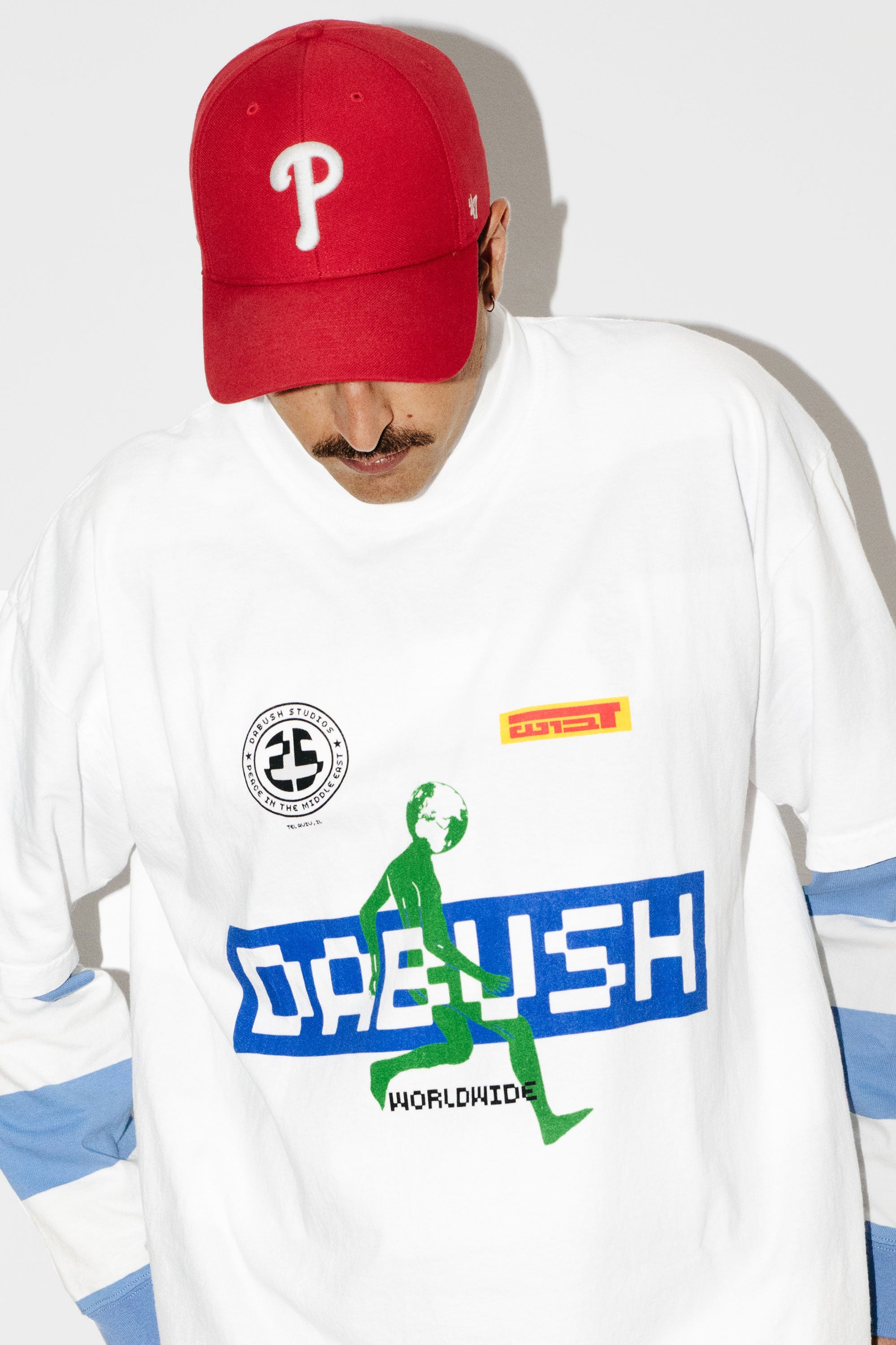 A model wearing an oversized DABUSH t-shirt in white, with colorful graphics, made from 100% cotton.

דוגמן לובש טישרט אוברסייז של דבוש בצבע לבן, עשויה 100% כותנה עם גרפיקה צבעונית.
