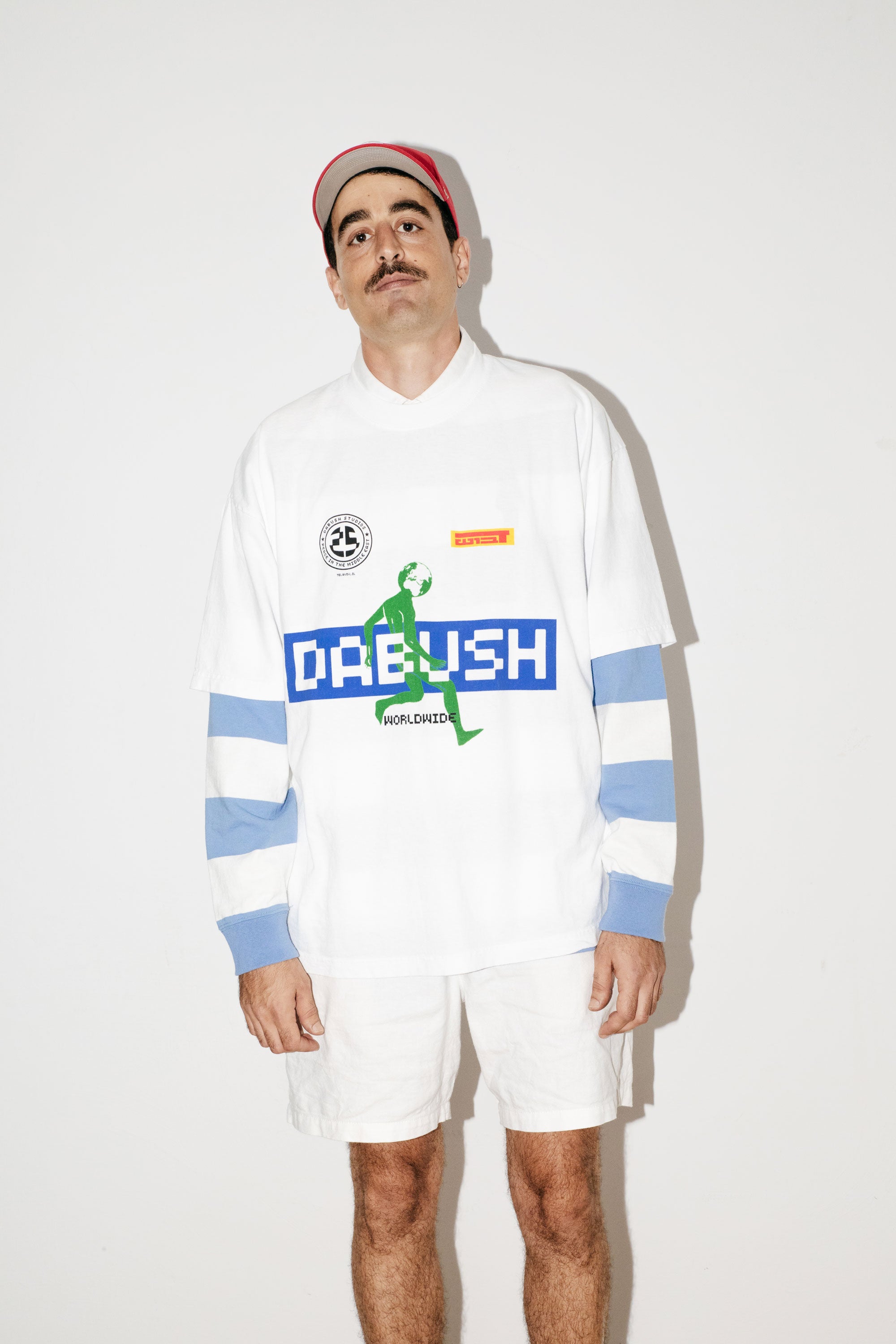 A model wearing an oversized DABUSH t-shirt in white, with colorful graphics, made from 100% cotton.

דוגמן לובש טישרט אוברסייז של דבוש בצבע לבן, עשויה 100% כותנה עם גרפיקה צבעונית.
